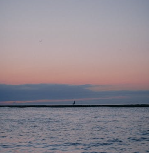 A serene sunset view over a calm body of water, with hues of pink and purple in the sky. A silhouette of a person is visible in the distance on a thin strip of land, adding a sense of solitude and tranquility to the scene.